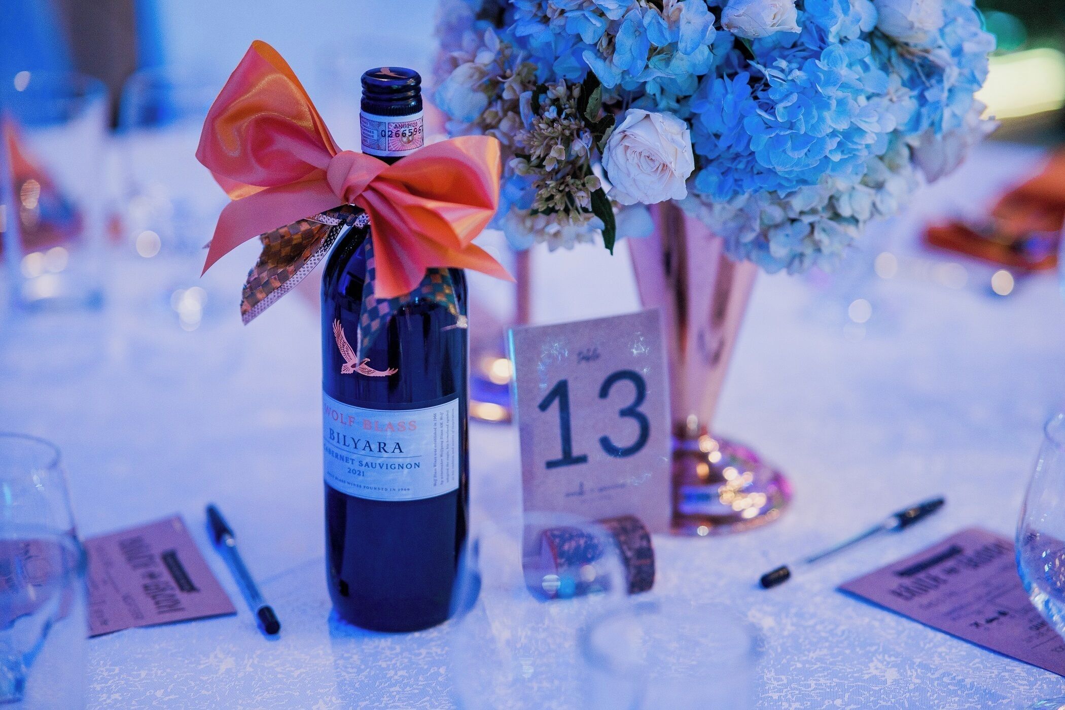 A bottle of wine with a bow, a number card, and a bouquet of flowers on a table.