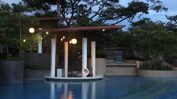 Zona de piscina con área de descanso cubierta, rodeada de árboles y un muro de piedra.