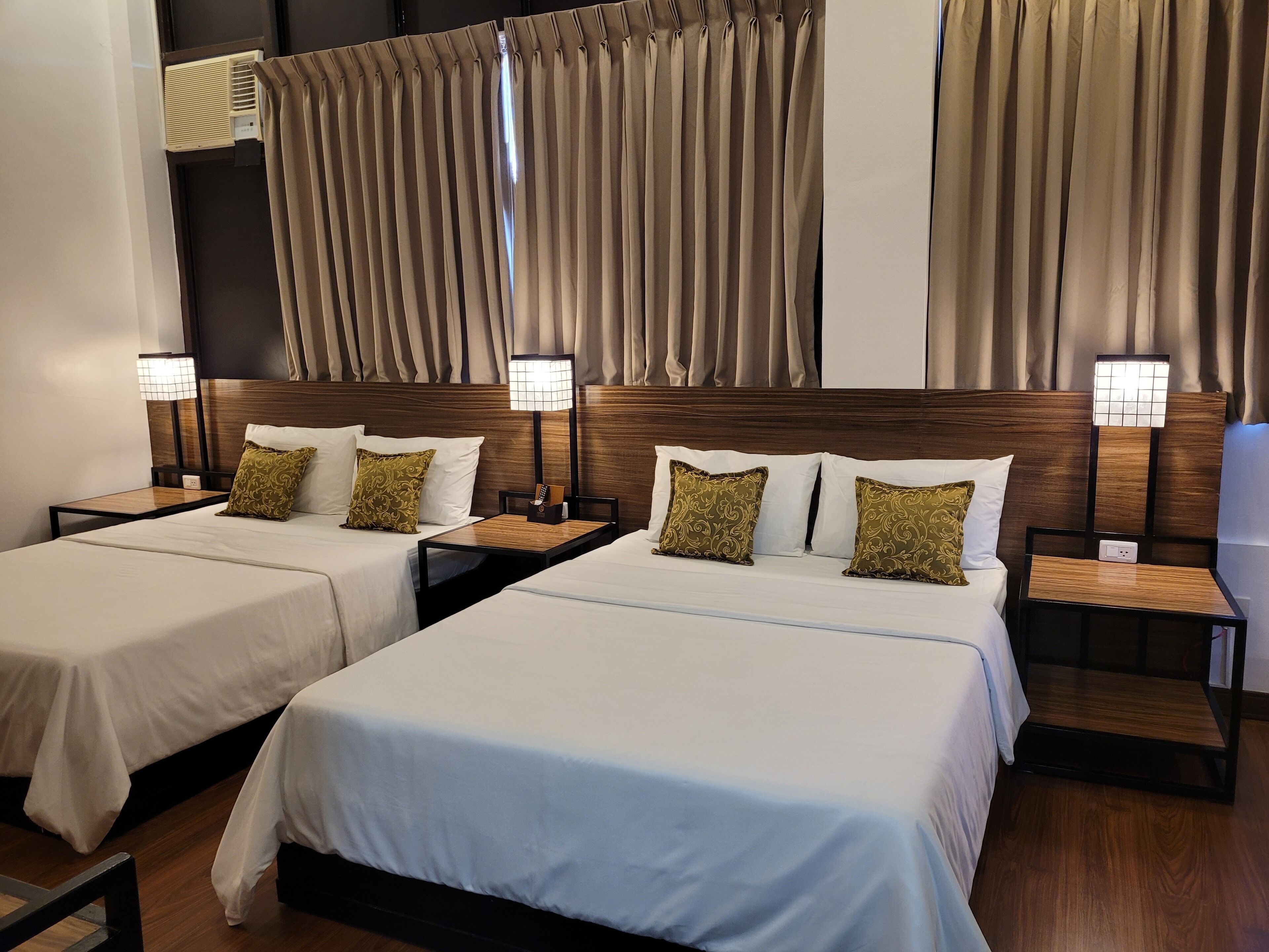 Een hotelkamer met twee bedden, elk met een houten hoofdeinde, wit beddengoed en decoratieve kussens. Er staan nachtkastjes met lampen en er is een wandgemonteerde airconditioning.