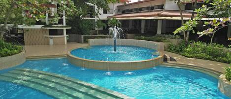 Une piscine dotée d’une fontaine centrale, entourée d’une terrasse carrelée et de verdure.
