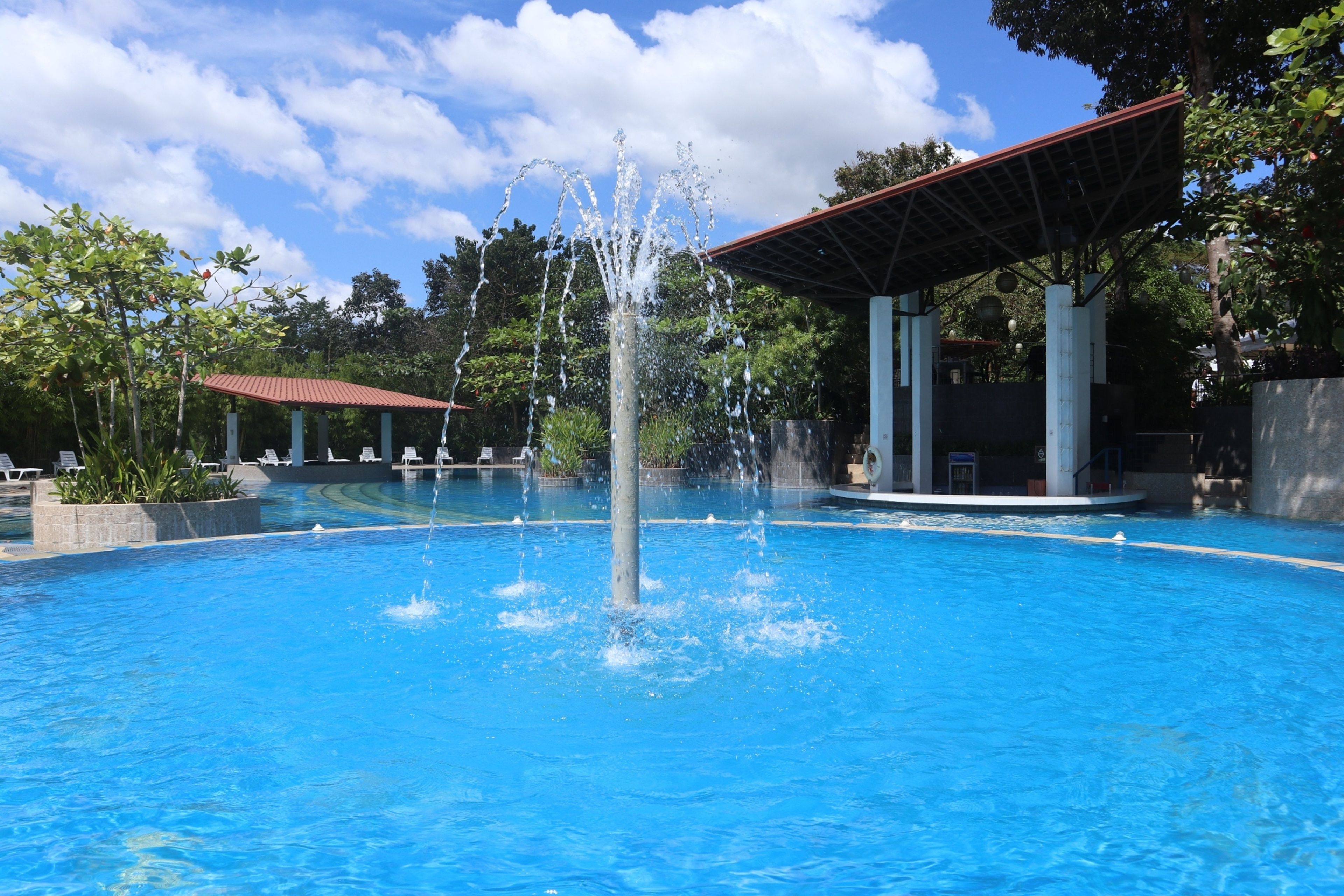 A swimming pool with a central fountain, surrounded by lounge chairs and a covered pavilion.