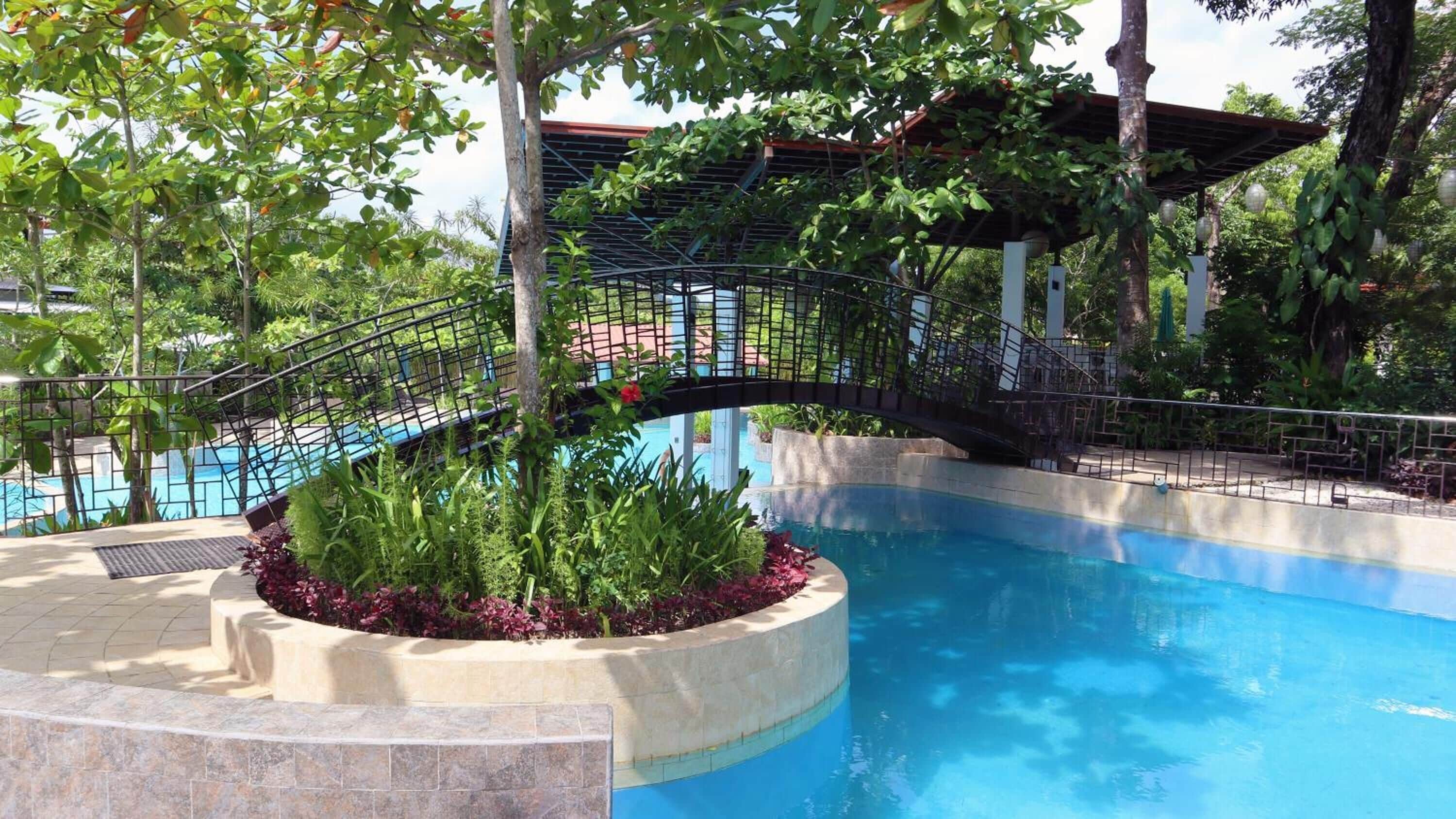 Una piscina con borde curvo, rodeada por una boveda de piedra y abundante vegetación. Un puente metálico cruza la piscina, conduciendo a un espacio cubierto con techo.