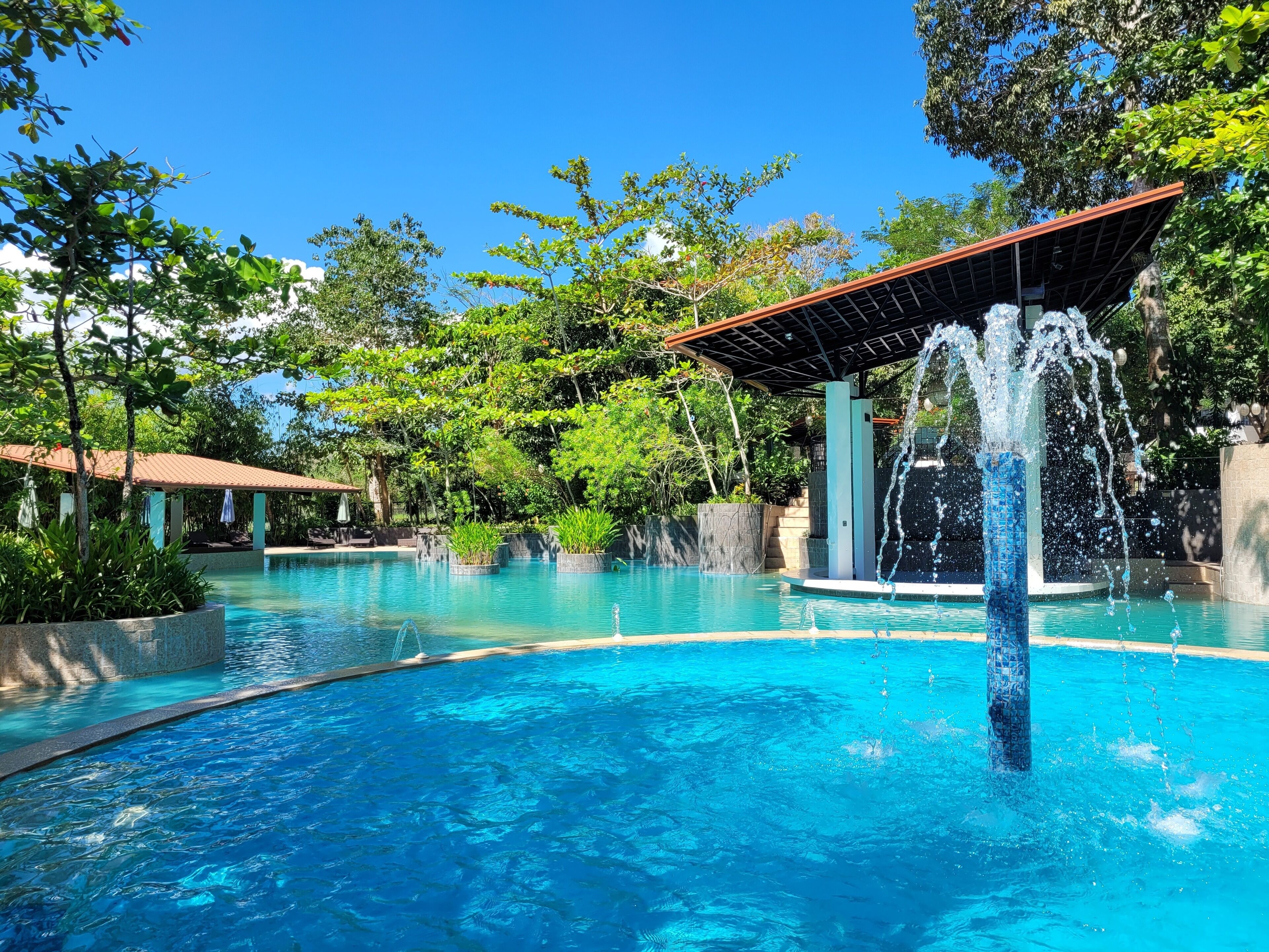 Una piscina d'un blau intens amb una font d'aigua, envoltada de vegetació exuberant i una zona de seients coberta.