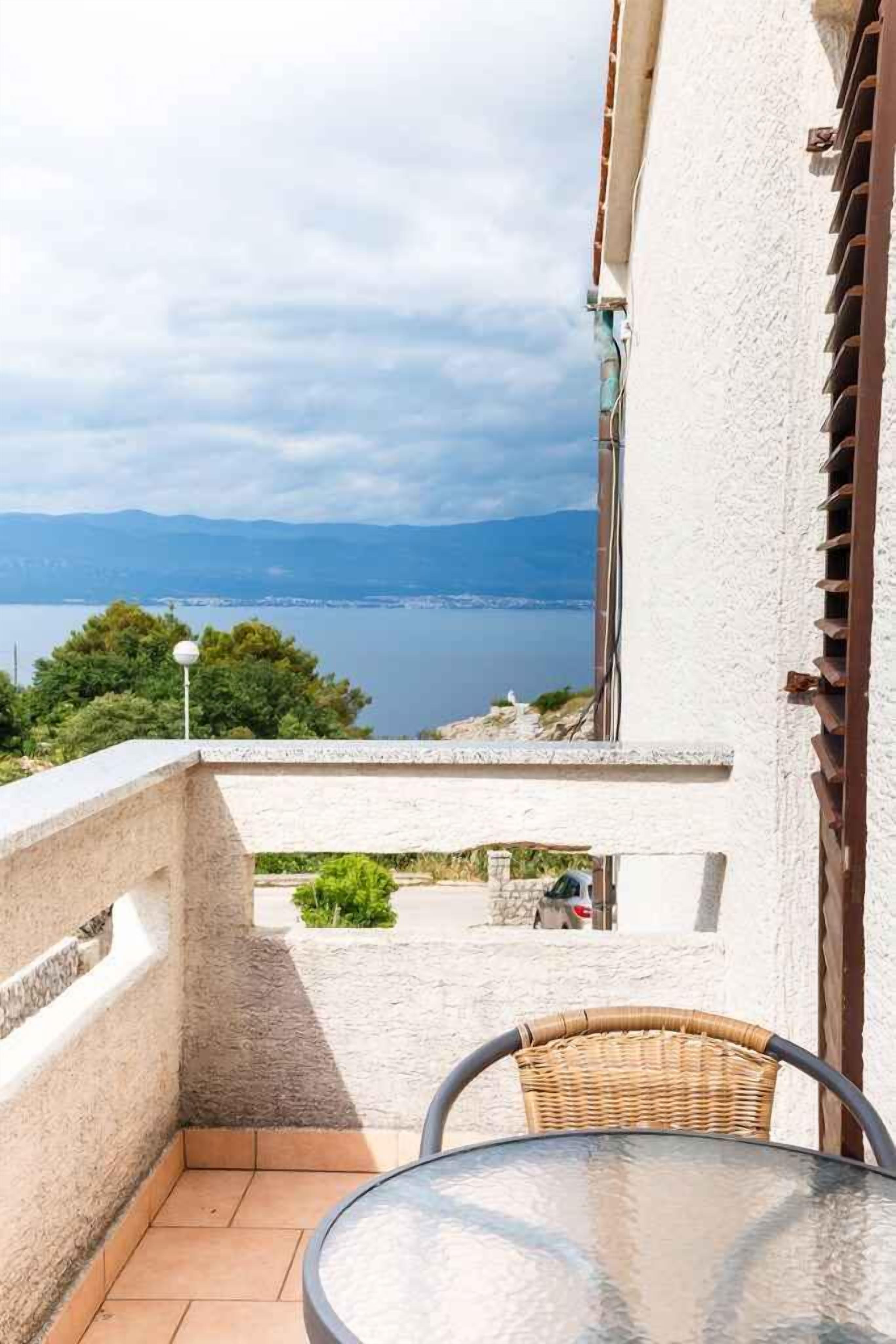 A balcony with a view of a lake and mountains, a wicker chair, and a table.