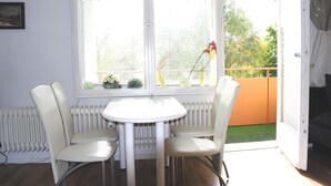 Een kamer met een witte tafel en vier stoelen, een radiator, een raam met kantgordijnen en uitzicht op een tuin en bomen.