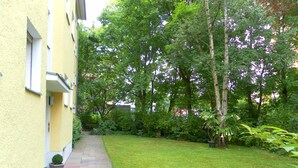 Een pad dat leidt naar een groen gazon met bomen en struiken, een geel gebouw met een raam, en een bloempot met plant.