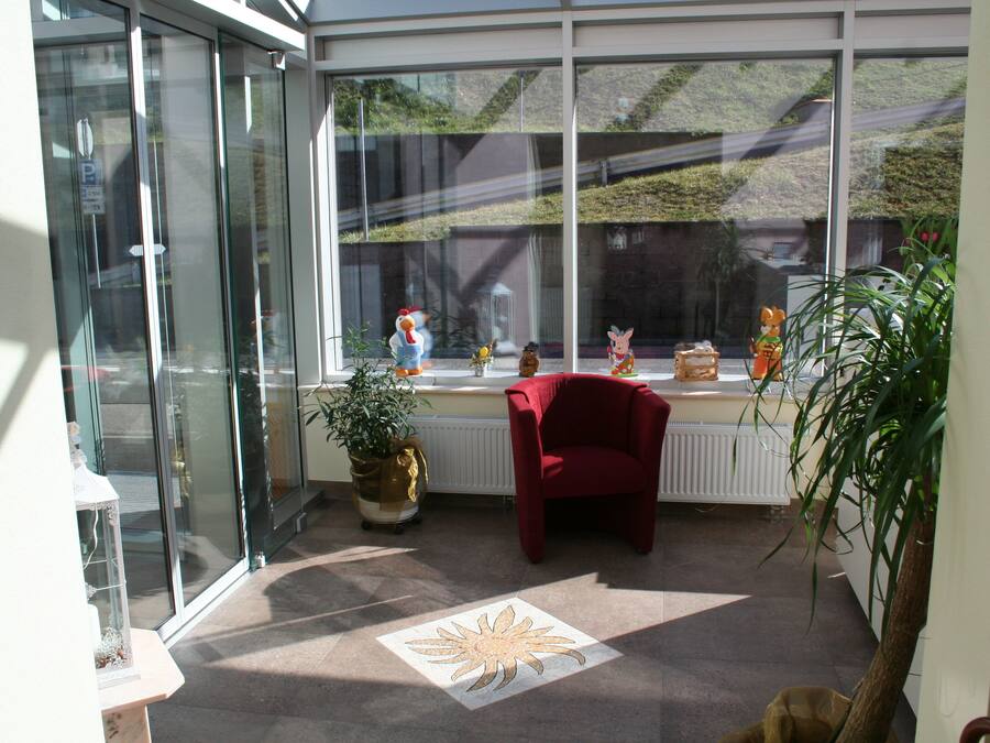 Una habitación iluminada por el sol, con una silla roja, una planta en maceta y una ventana que ofrece vistas a un edificio y vegetación.