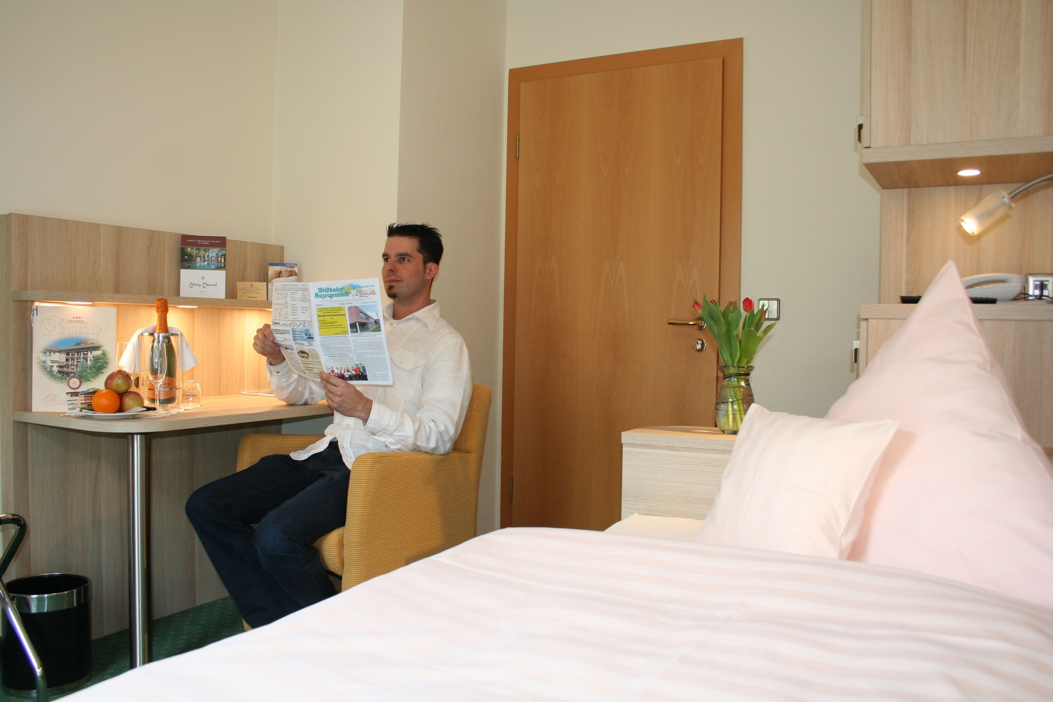 Un hombre leyendo una revista en una habitación de hotel con una cama, un escritorio y una pequeña mesa con frutas y una botella.
