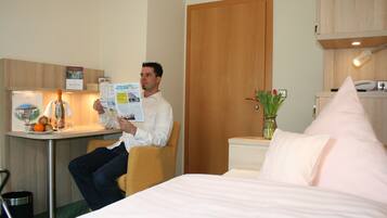 Un hombre leyendo una revista en una habitación de hotel con una cama, un escritorio y una pequeña mesa con frutas y una botella.