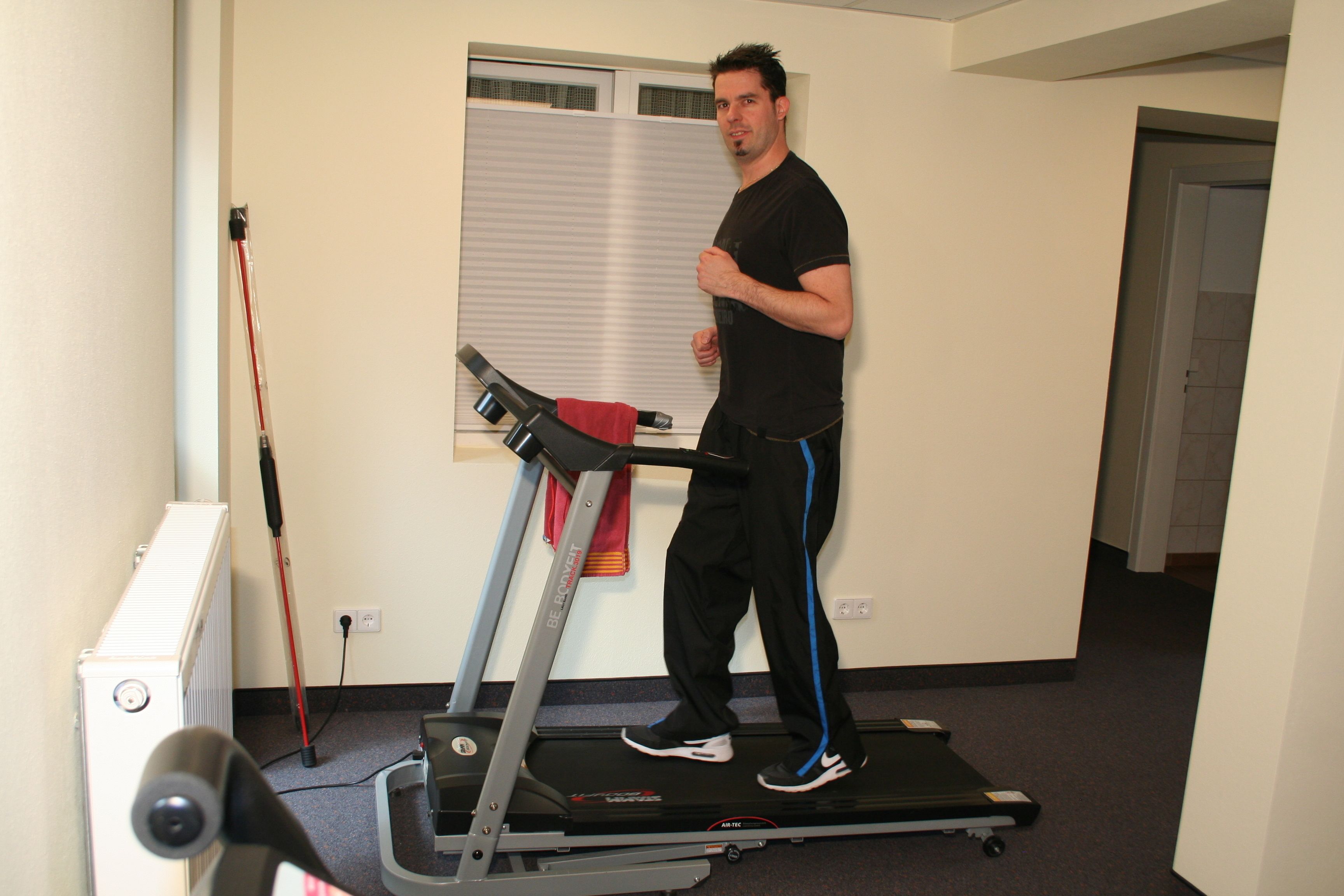 Un homme utilise un tapis de course dans une pièce où se trouvent un radiateur, une fenêtre avec des stores et une porte au fond.