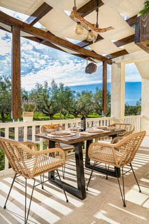 Een terras met een tafel voor vier personen, omringd door rieten stoelen, onder een houten pergola met uitzicht op olijfbomen en de zee.