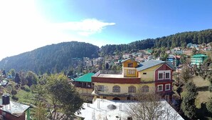 A hillside village with colorful buildings, a prominent yellow and red house, and a dense forested mountain in the background. - Mountain And Moon (Dharamshala)