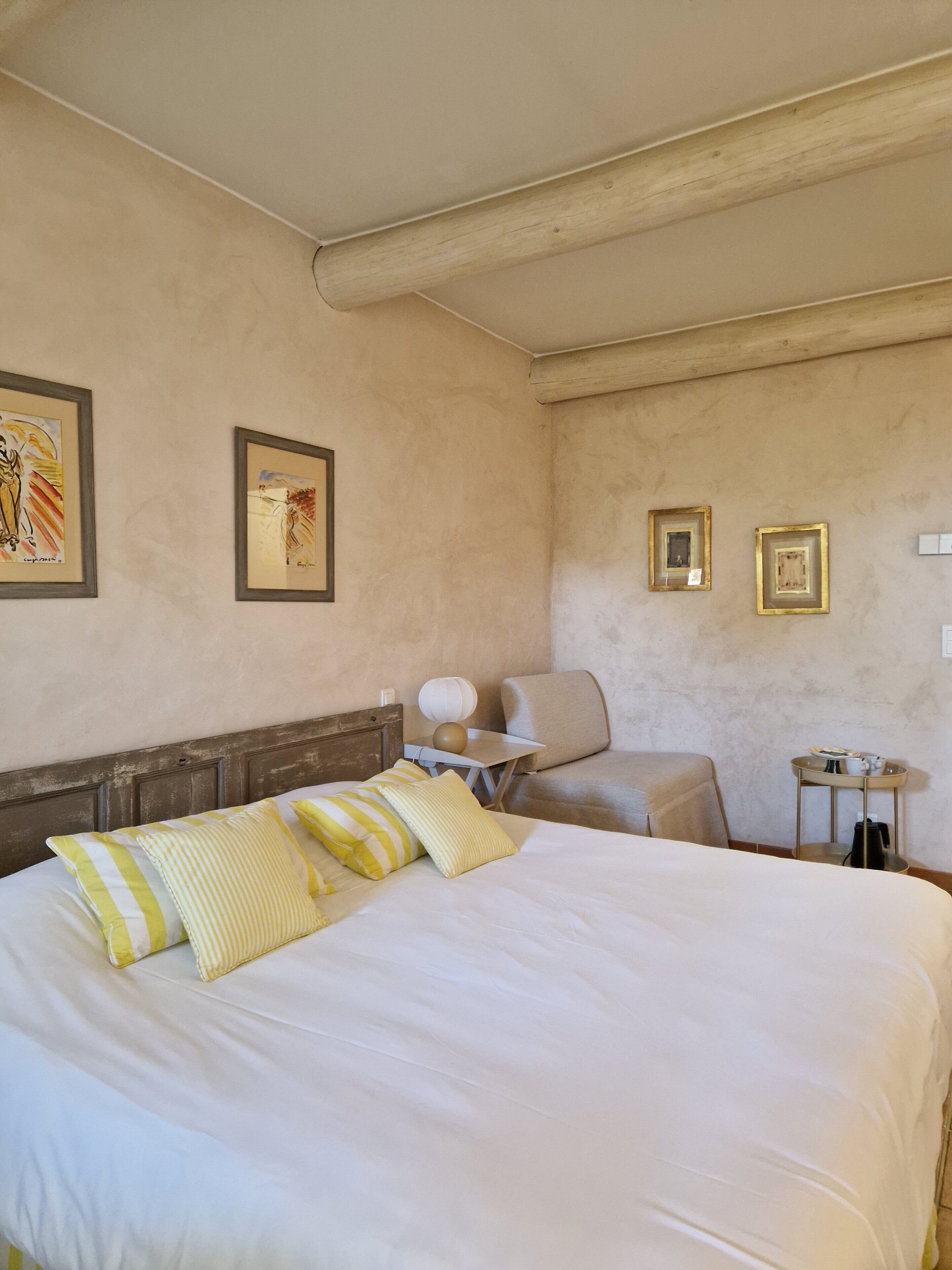 A hotel room with a large bed, a small table, a chair, and two framed pictures on the wall.