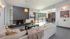 A modern living room with a fireplace, a dining area, and a large window offering a view of the outdoors. - Filerimos Villa (Rhodes)