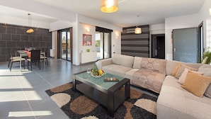 A modern living room with a glass coffee table, a beige sofa, and a dining area with a table and chairs. - Filerimos Villa (Rhodes)