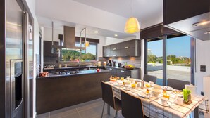 A modern kitchen with a central island, large windows, and a dining area set for breakfast. - Filerimos Villa (Rhodes)
