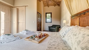Un dormitorio con cama, cabecera de madera, ventana, silla y bandeja con toallas y productos para el desayuno.