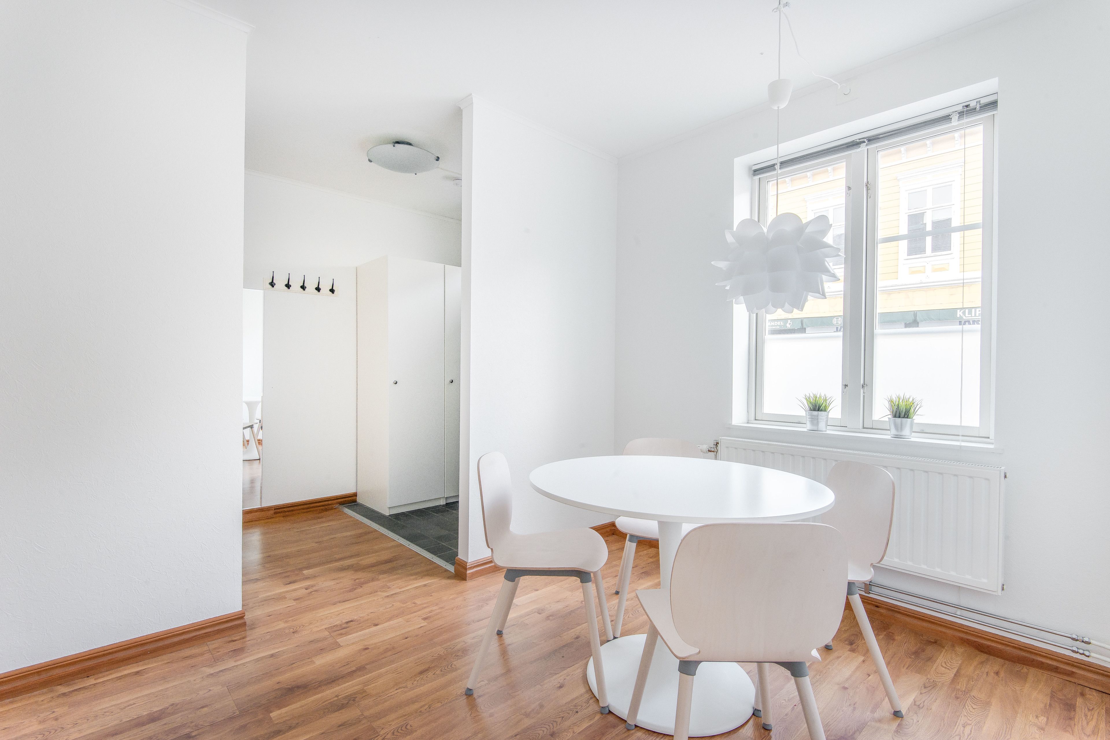 Una habitación moderna y luminosa con una mesa redonda blanca y sillas a juego, un ventanal con persianas y vistas a la calle.