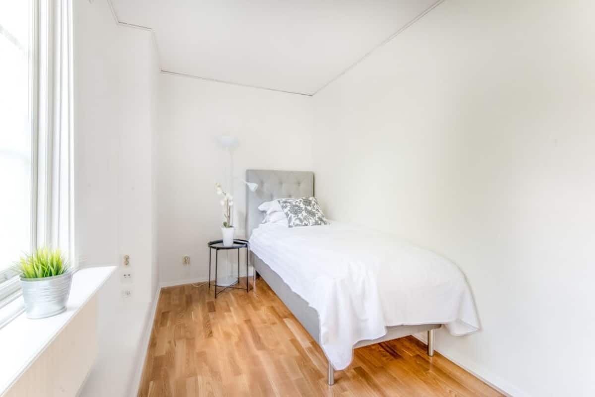 A bedroom with a bed, a bedside table, a lamp, and a window with a plant.