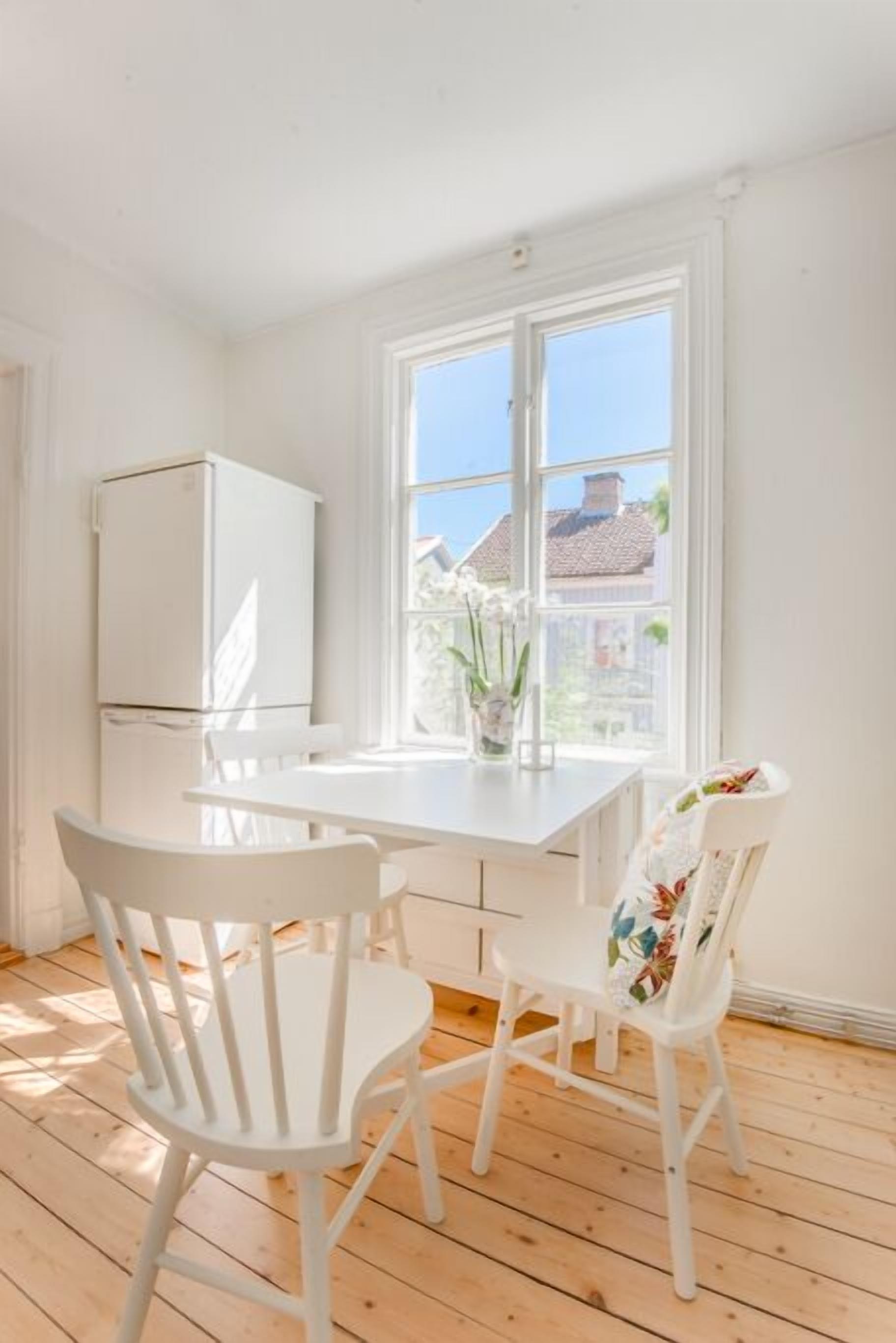 A bright room with a white table and chairs, a window with a view of houses, and a white refrigerator.