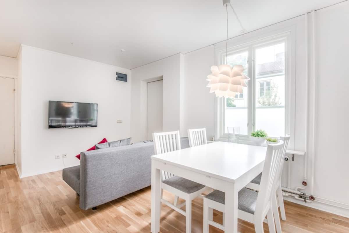 A modern living room with a white dining table, grey sofa, and a TV mounted on the wall.