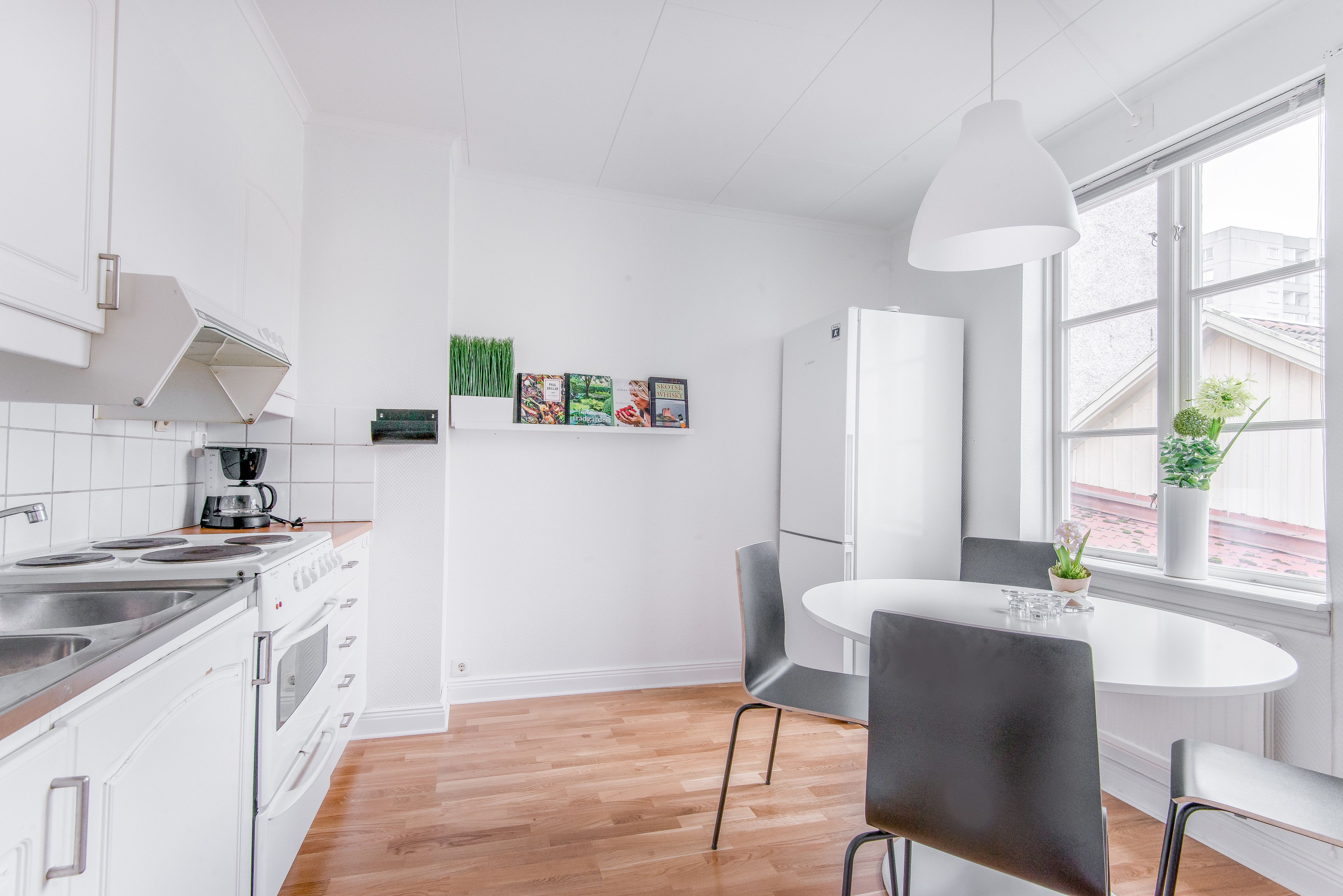 Una cocina moderna con armarios blancos, una estufa y una cafetera. Hay una zona de comedor con una mesa y sillas blancas.