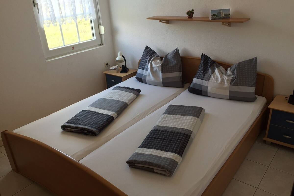 A double bed with two pillows and two folded blankets. There is a nightstand with a lamp and a small shelf above the bed.