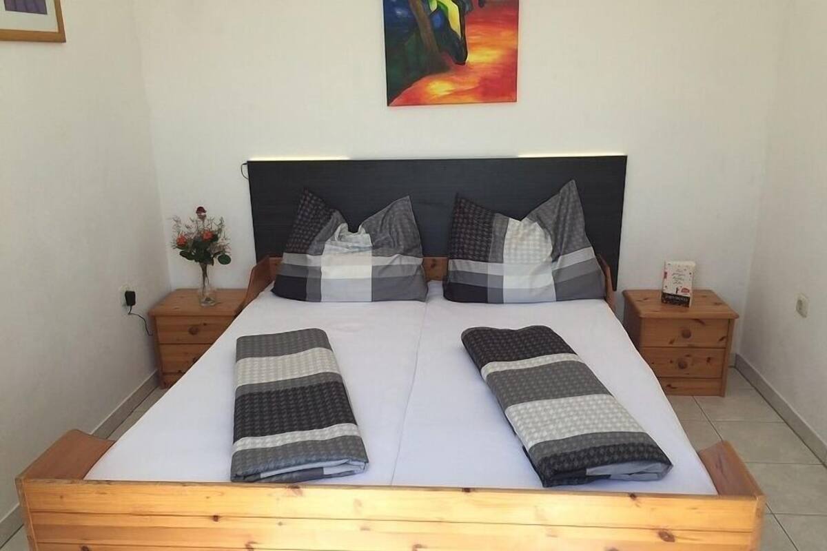 A double bed with a wooden frame, two nightstands with drawers, a vase with flowers, and a painting on the wall.