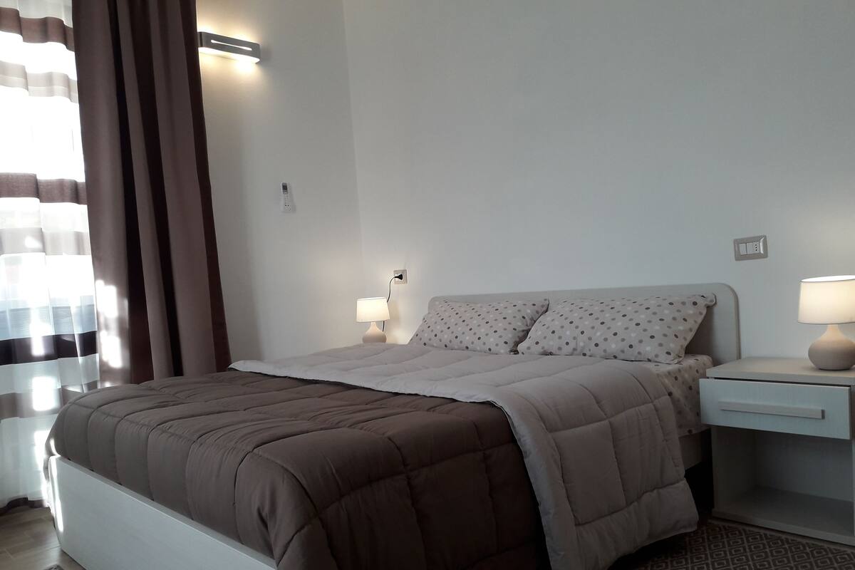 Un lit bien fait, avec une couette marron, une table de chevet surmontée d’une lampe et un tapis à motifs sur le sol.