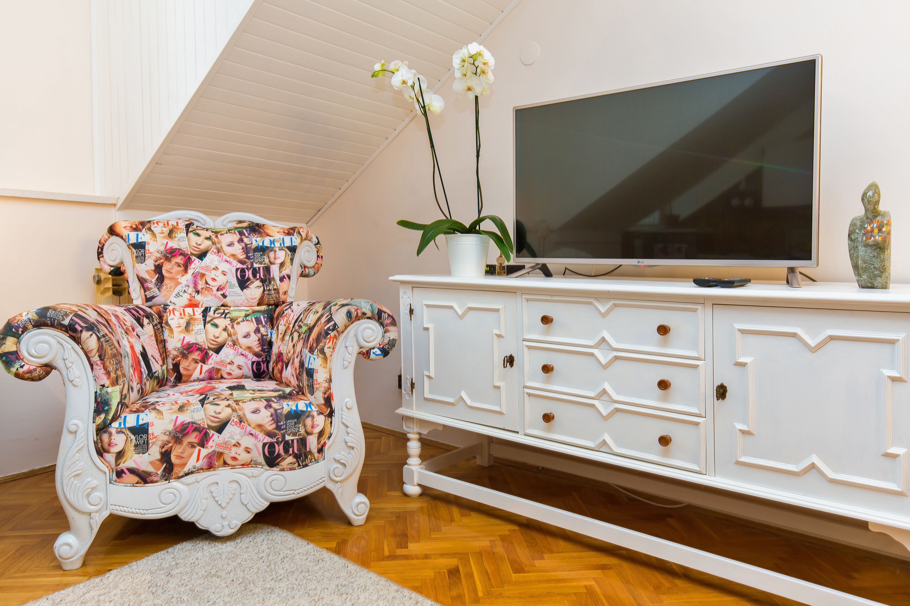 Un soggiorno con una poltrona bianca ricoperta di riviste, un mobile bianco con cassetti, una televisione a schermo piatto e una pianta di orchidea.