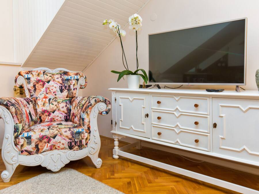 Un soggiorno con una poltrona bianca ricoperta di riviste, un mobile bianco con cassetti, una televisione a schermo piatto e una pianta di orchidea.