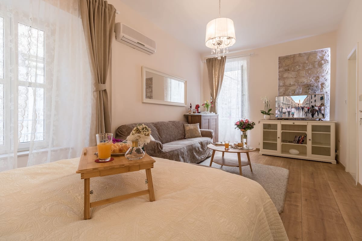 Quarto de hotel acolhedor com uma cama grande, uma mesa de cabeceira de madeira com um copo de sumo de laranja, uma pequena mesa redonda com um vaso de flores, um sofá, um armário com livros e uma janela grande com cortinas.