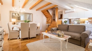 Una sala de estar acogedora con techo de madera, un comedor con sillas blancas, un sofá marrón y una cocina al fondo.