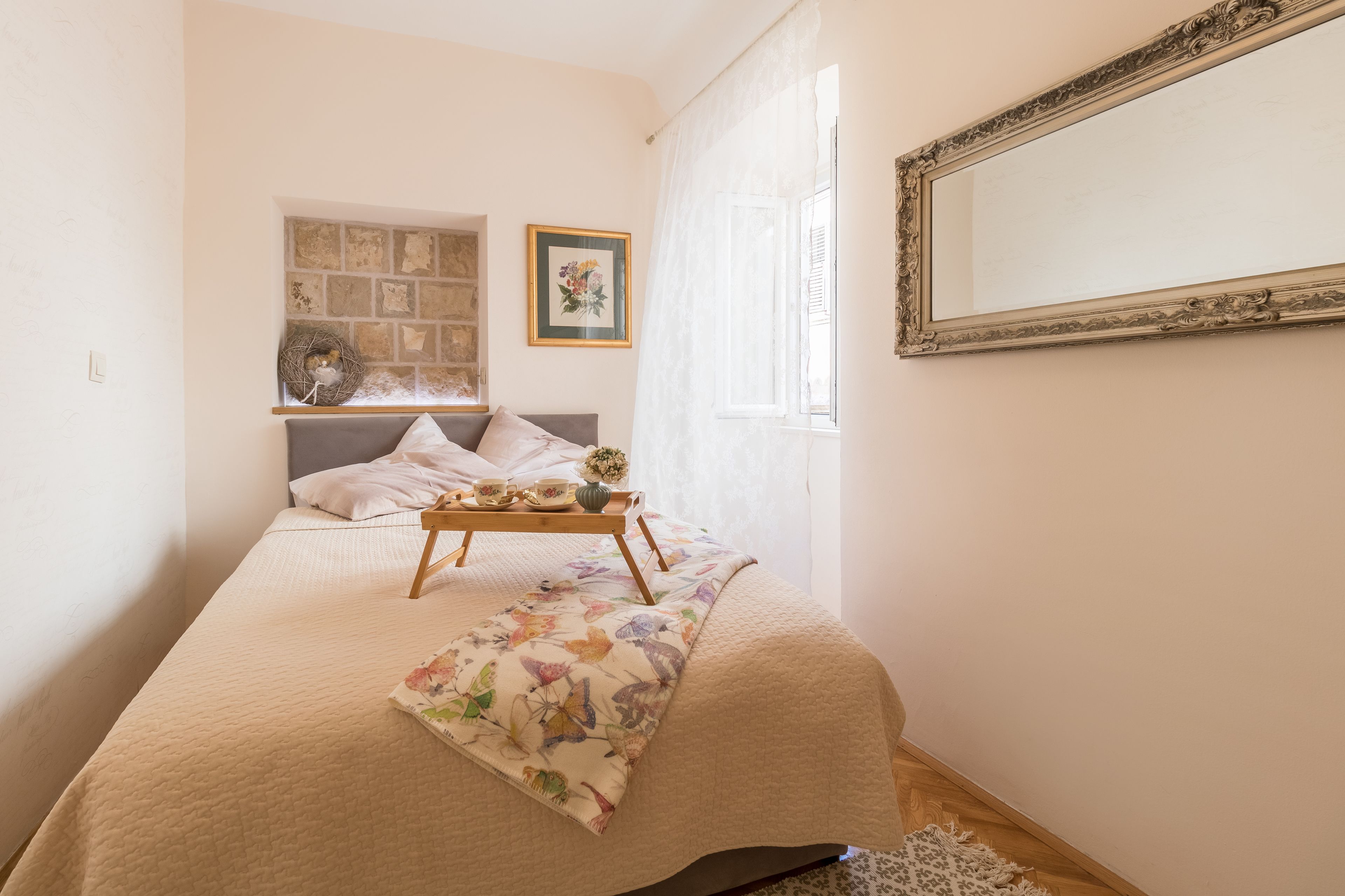 Ein Schlafzimmer mit einem Bett, einem Nachttisch mit Tablett, einem gerahmten Bild, einem Fenster mit Vorhängen und einem Spiegel an der Wand.