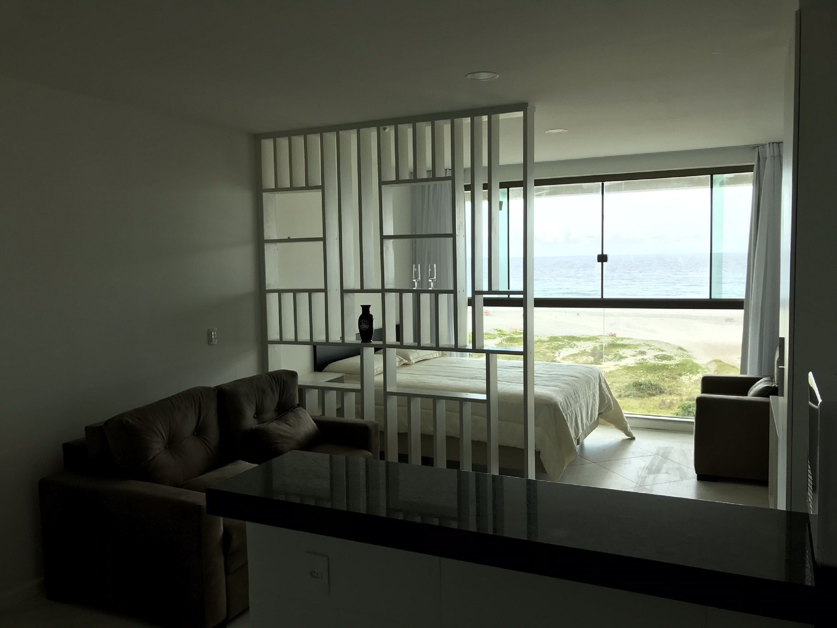 A modern hotel room with a large window offering a view of the beach, a bed, a sofa, and a small table.