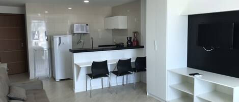 A modern kitchen with white cabinets, a microwave, and a coffee maker. There is a dining area with black chairs and a white table. A TV is mounted on the wall, and a sofa is in the living area.