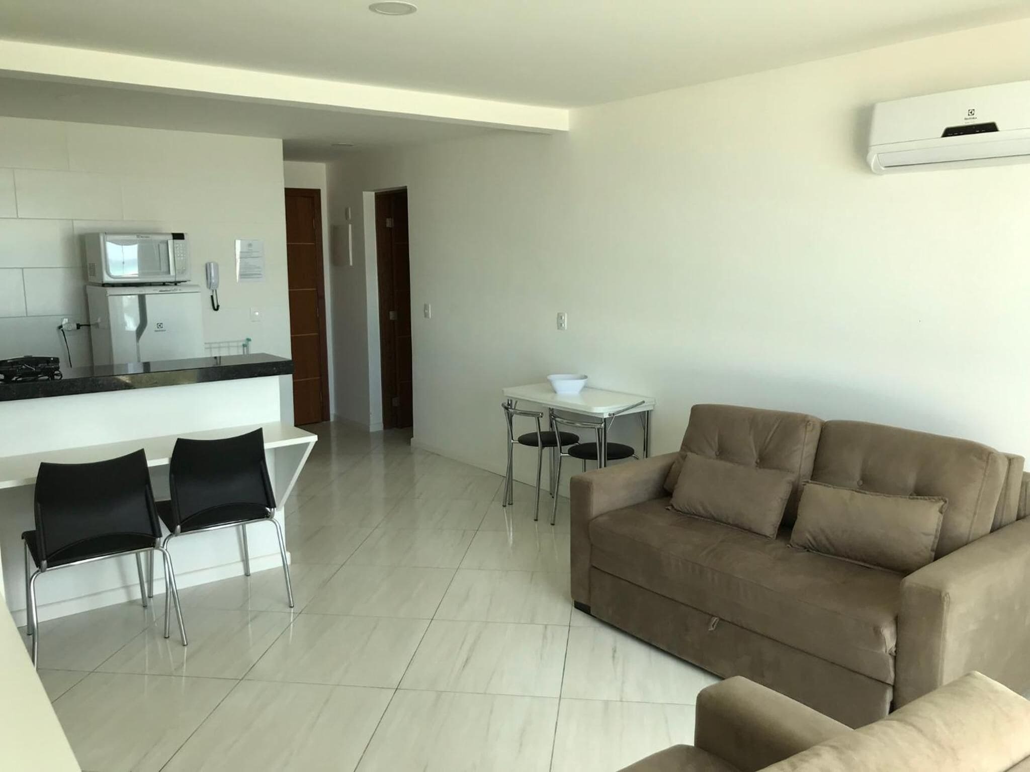 A modern living room with a beige sofa, a dining area with two chairs, a kitchen with a microwave, and a small table with a bowl.