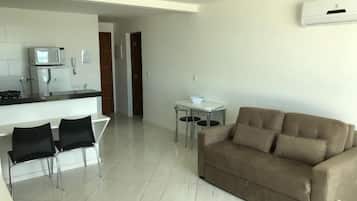 Uma sala moderna com um sofá bege, um espaço de jantar com duas cadeiras, uma cozinha com micro-ondas e uma pequena mesa com uma tigela.