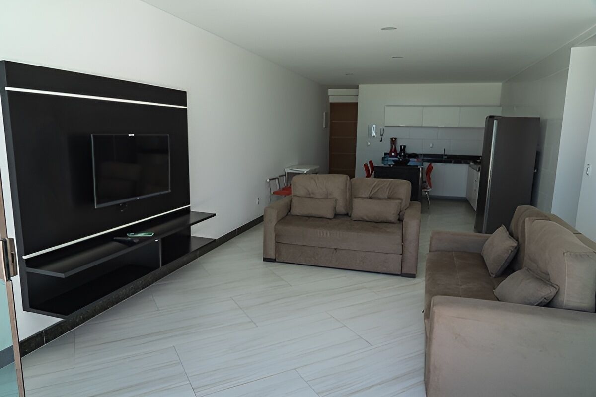 Una sala de estar moderna con un televisor de pantalla plana, dos sofás color beige, una mesita de centro y una cocina al fondo.