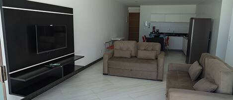 Una sala de estar moderna con un televisor de pantalla plana, dos sofás color beige, una mesita de centro y una cocina al fondo.