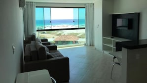 A modern living room with a large window offering a view of the sea and a distant building. - Sophia II Residence Inn (Arraial do Cabo)