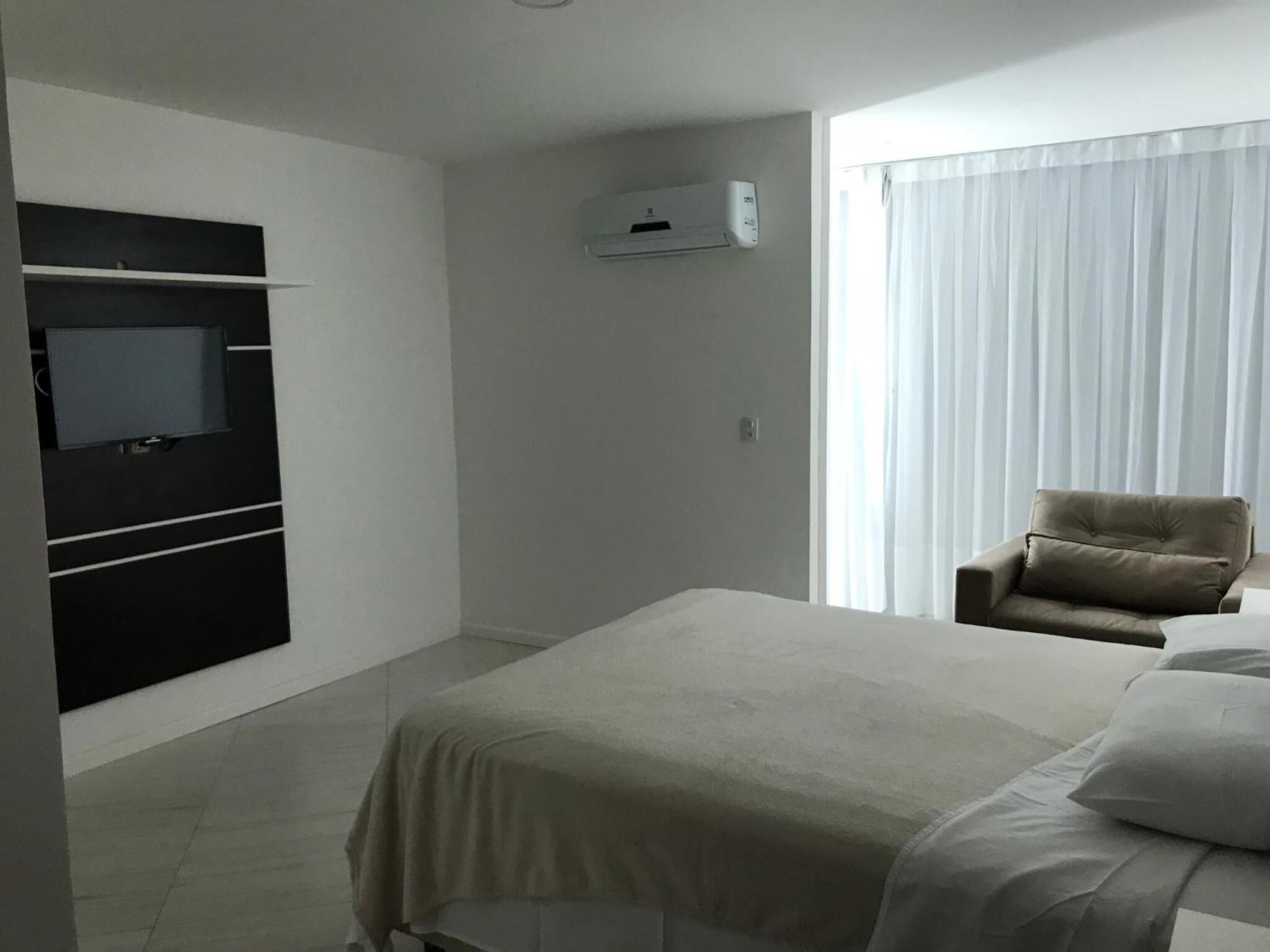 Una habitación de hotel con una cama, un televisor montado en un panel negro, un equipo de aire acondicionado, una ventana con cortinas traslúcidas y una silla.