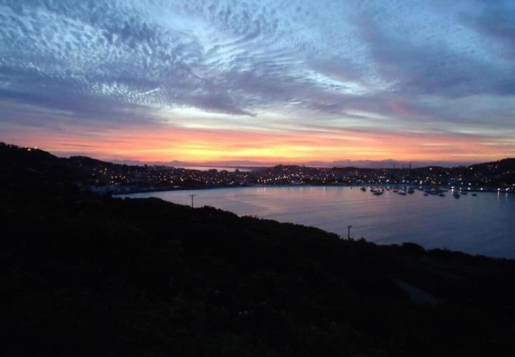 Un paisaje urbano costero al atardecer, con la línea de la costa a la vista, una masa de agua y luces de la ciudad en la distancia.