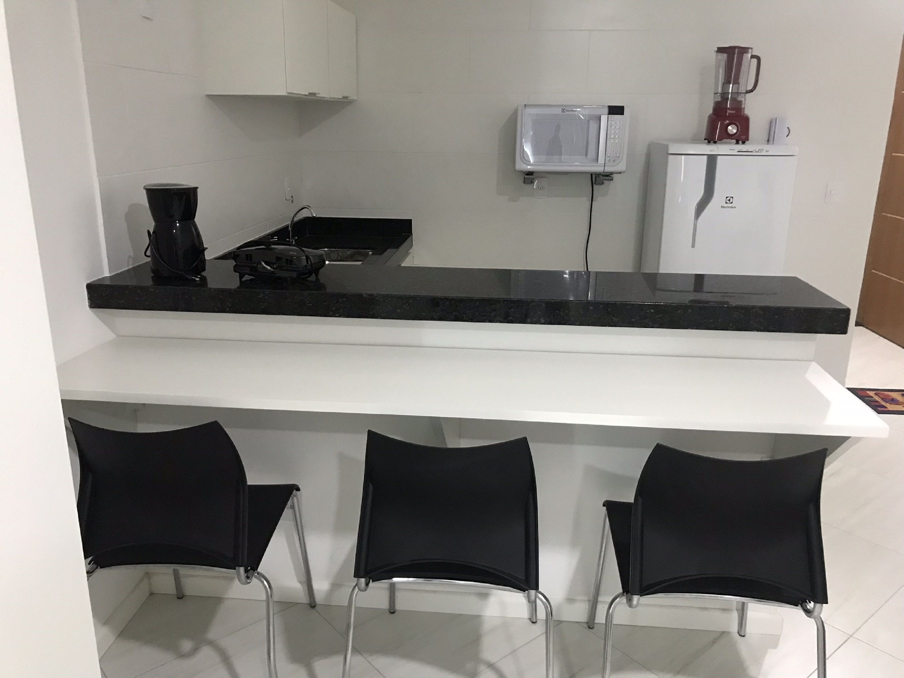 A kitchen with a black countertop, white cabinets, a microwave, a blender, and three black chairs.