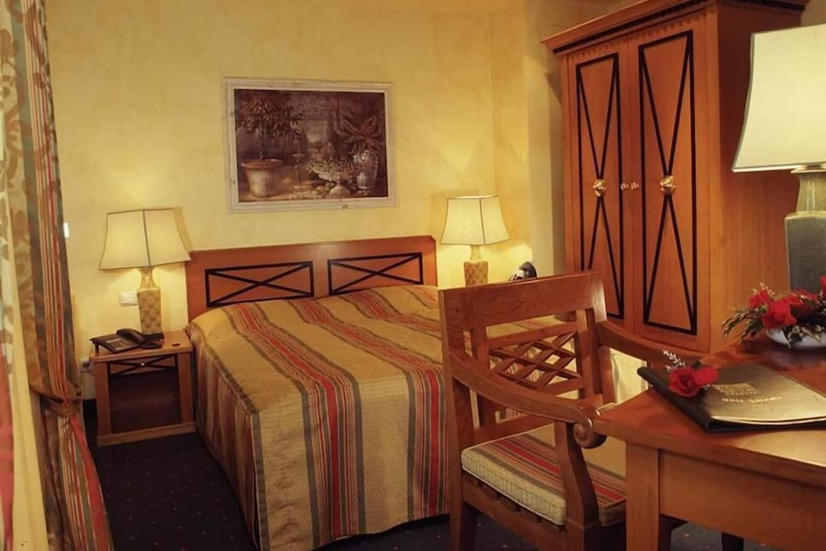 Quarto de hotel com cama, duas luminárias laterais, uma cadeira de madeira, uma escrivaninha com luminária e um quadro na parede.