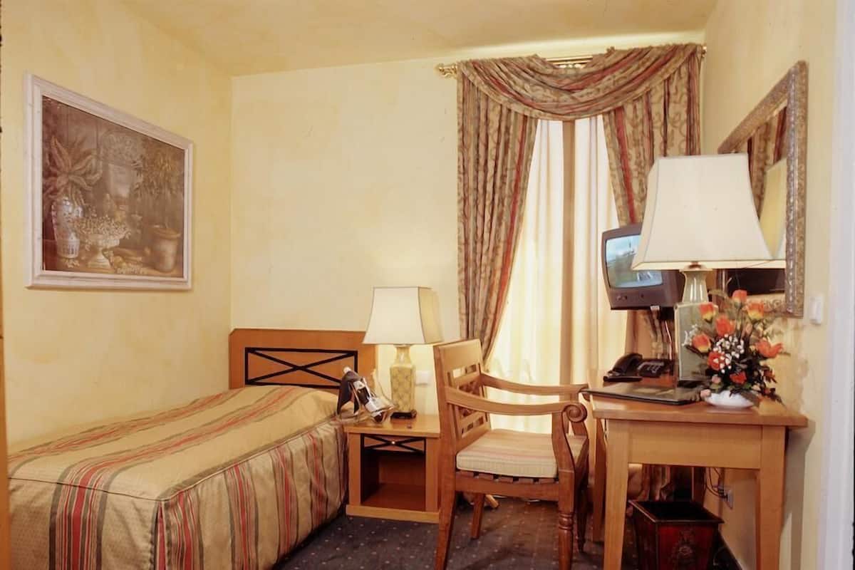 Quarto de hotel com uma cama de solteiro, uma mesa com cadeira, telefone, abajur e um quadro na parede.