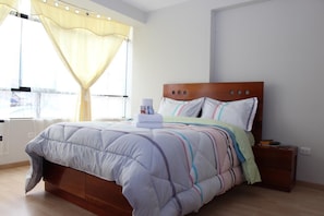 A neatly made bed with a wooden headboard, a bedside table with a book and a remote control, and a window with sheer curtains. - Asiriq Inn Cusco (Cusco)