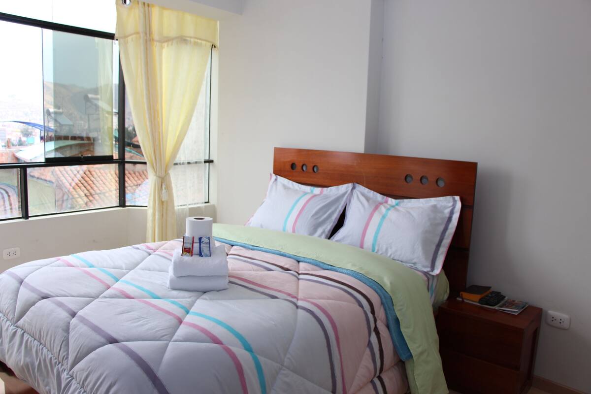 A neatly made bed with striped bedding, a wooden headboard, and a nightstand with a book and a remote control.