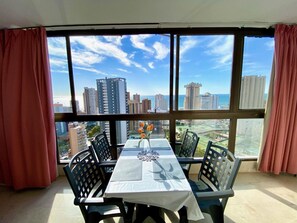 Un comedor con mesa y sillas, ventanas amplias con vista a la ciudad y cortinas rojas.