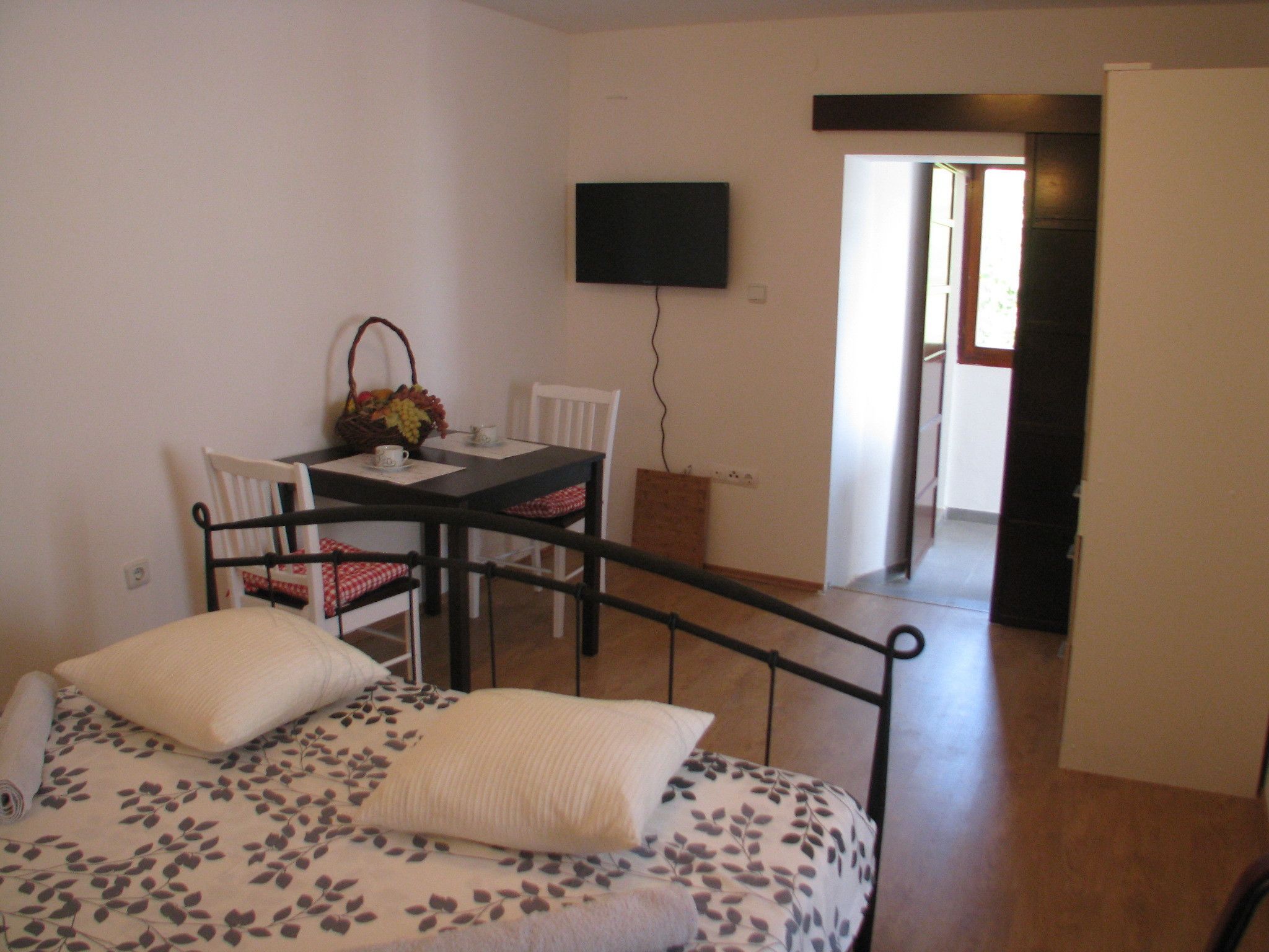 A bedroom with a bed, a small table with chairs, a TV mounted on the wall, and a door leading to another room.